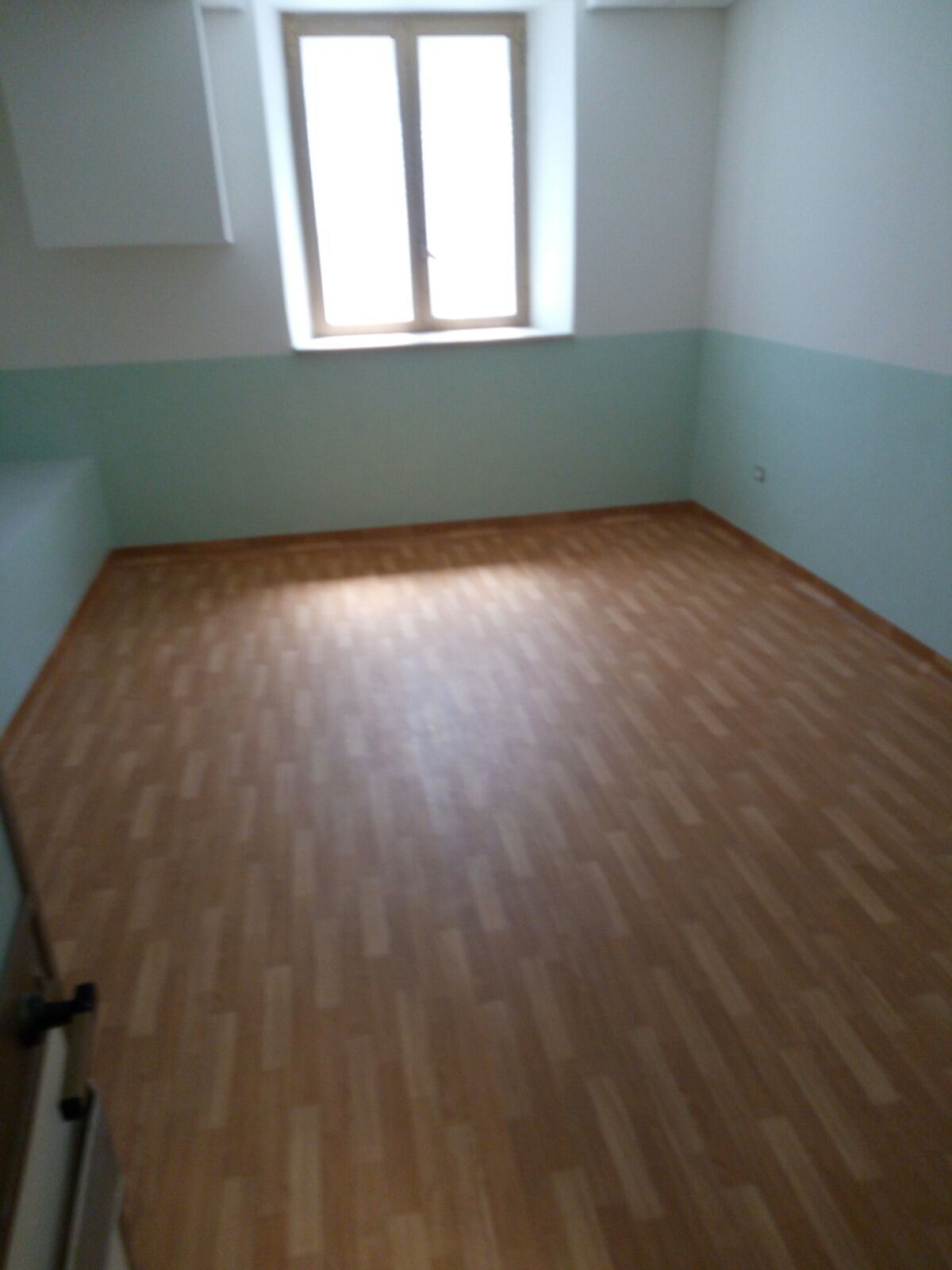 Stanza vuota con pavimento effetto legno chiaro, finestra e pareti verde chiaro e bianche.