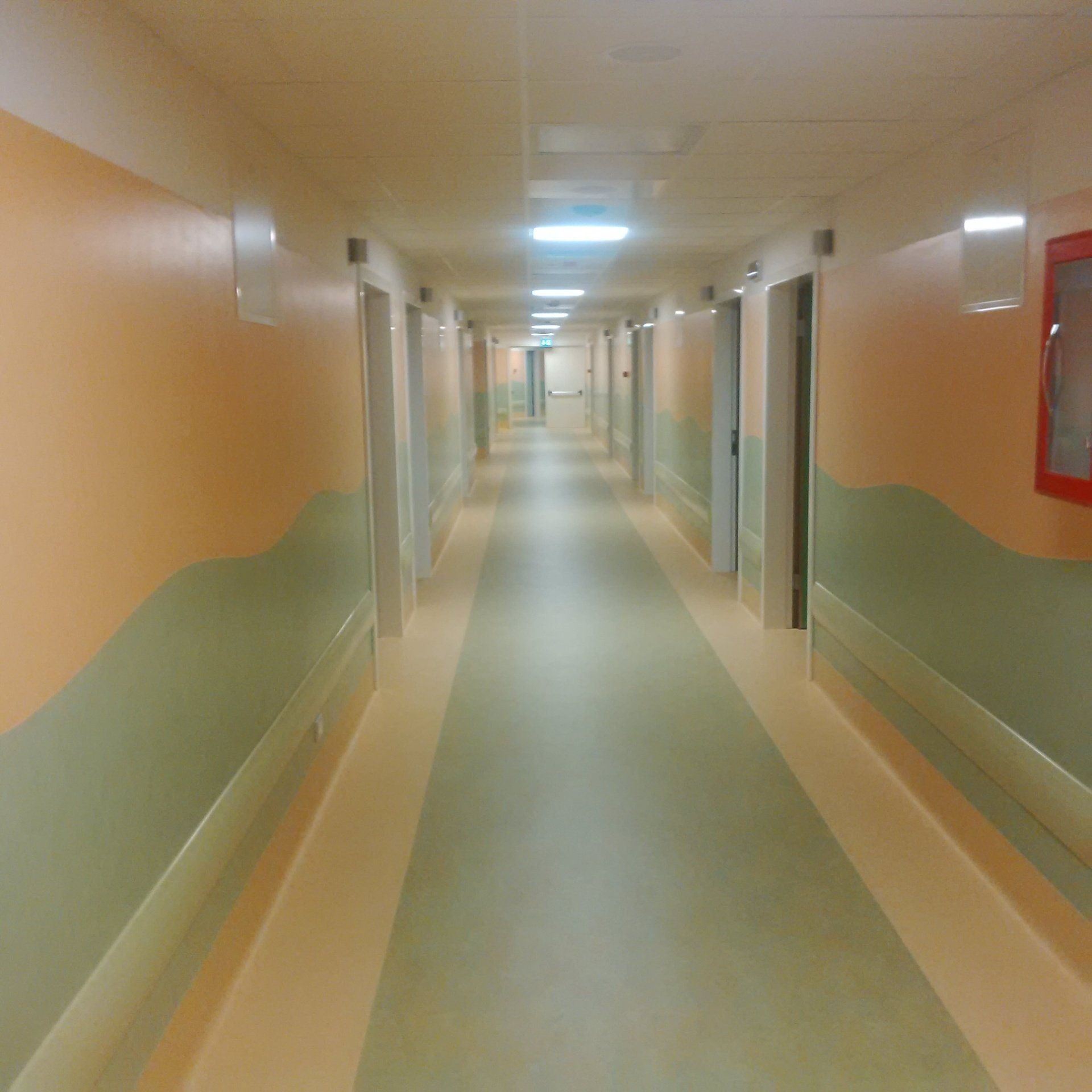 Lungo corridoio dell'ospedale con pareti, porte e luci arancioni e verdi.