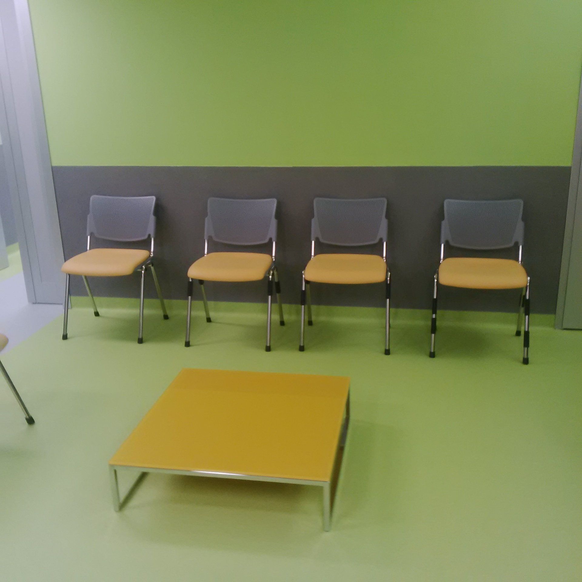 Quattro sedie con seduta gialla di fronte a un tavolino da caffè giallo in una sala d'attesa verde e grigia.