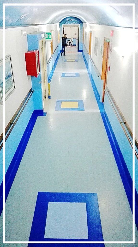 Lungo corridoio blu e bianco; una persona è in piedi in fondo. Ai lati sono visibili attrezzature antincendio, porte e opere d'arte.