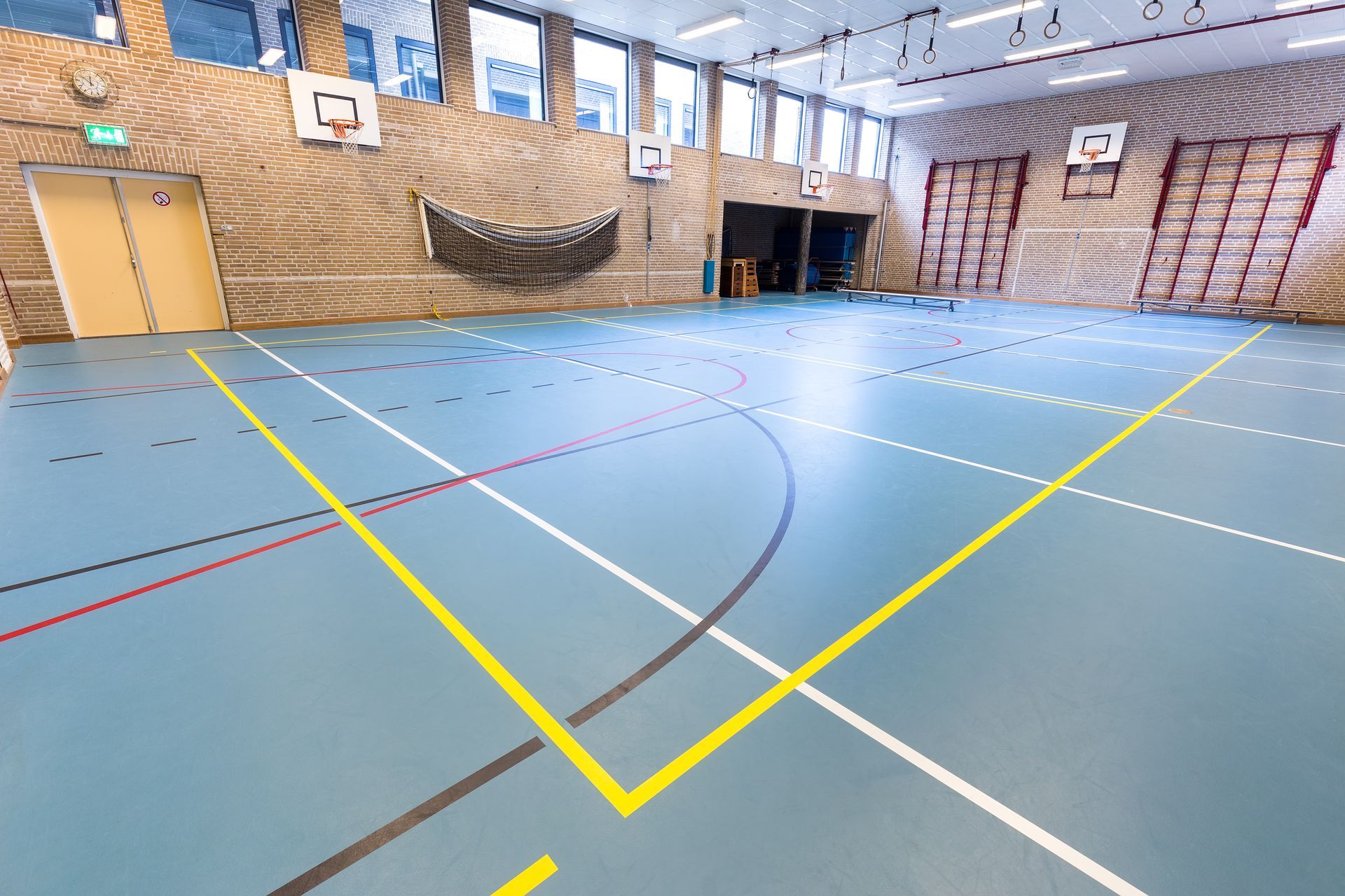 Interno della palestra con pavimento blu contrassegnato per gli sport, canestri da basket e parete in legno.