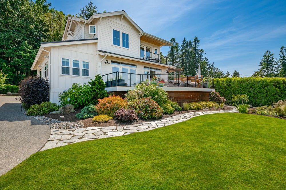 White house with a deck, lush green lawn, colorful shrubs, stone walkway, and sunny blue sky.