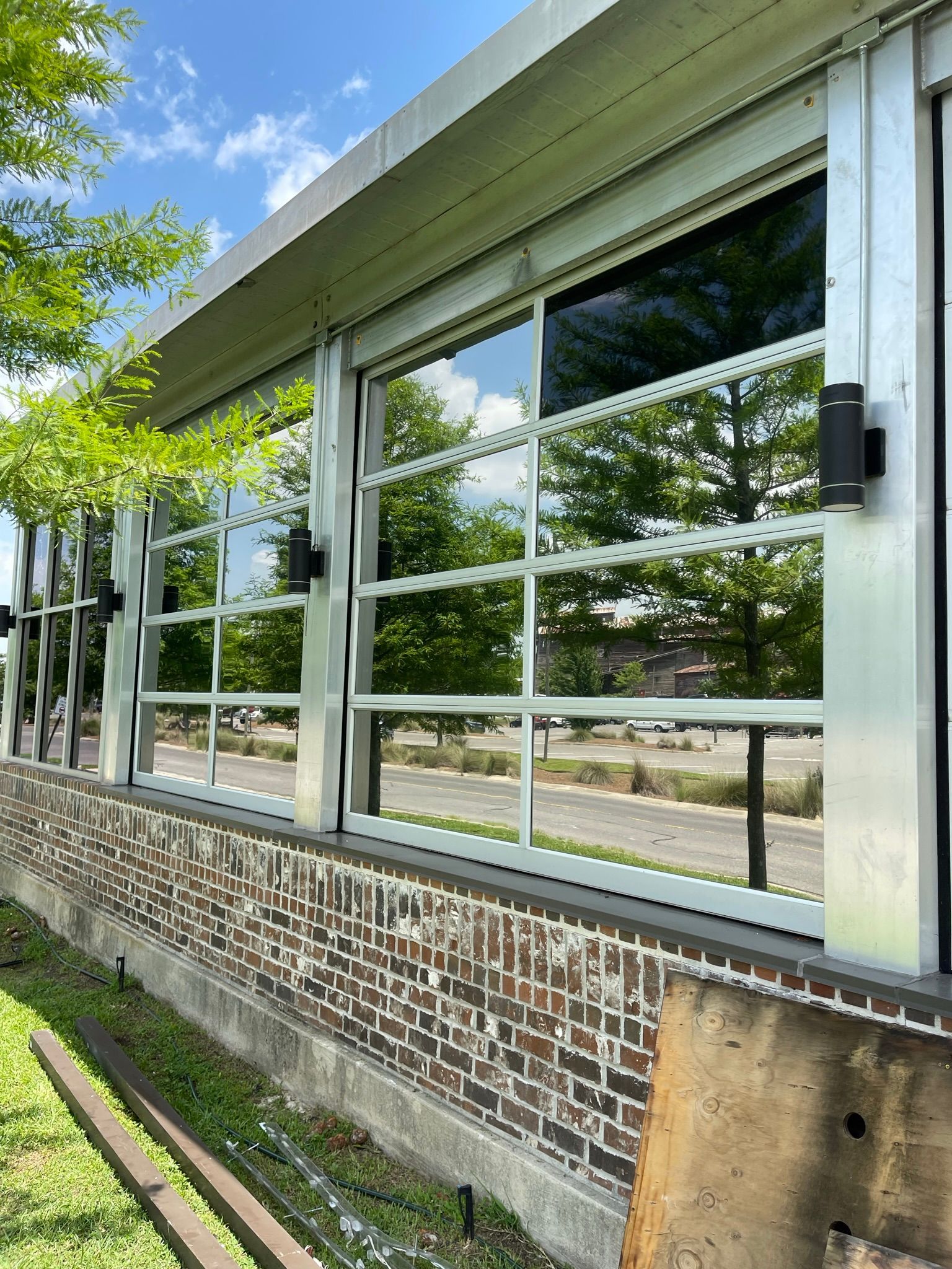 Glass wall with a reflective outdoor view. Brick base, greenery in the foreground, blue sky.