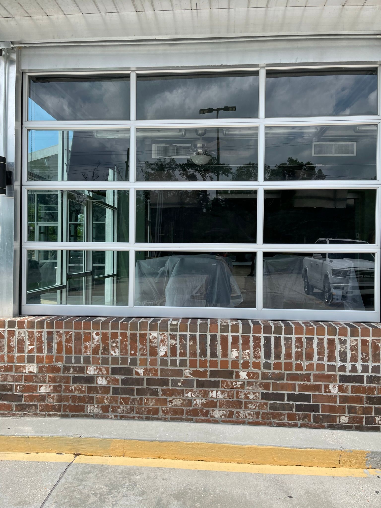 Glass garage door with white frame above a brick wall, reflecting trees and vehicles.