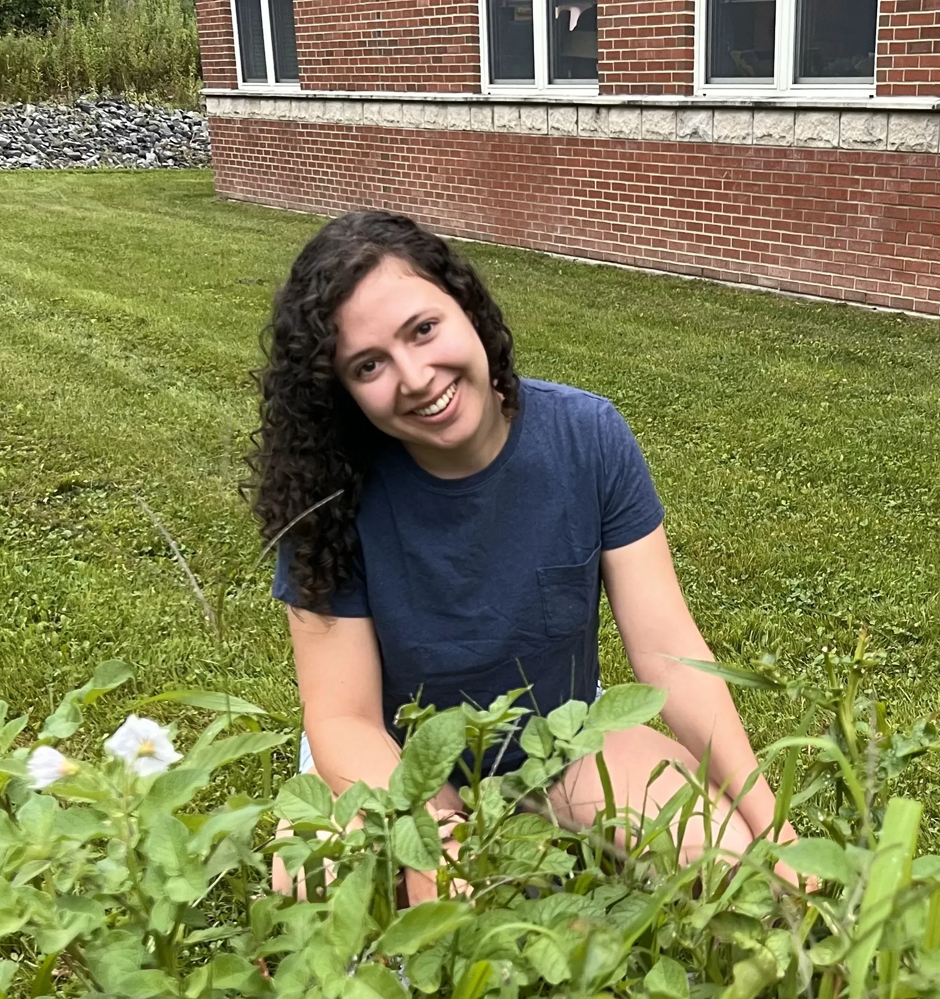Person smiling and sitting in grass with plants in front of a brick building