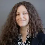 Woman with long, curly brown hair smiles, wearing a patterned blouse and dark blazer, against a gray background.