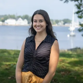 Smiling woman in a black top standing by a waterfront with boats and trees in the background