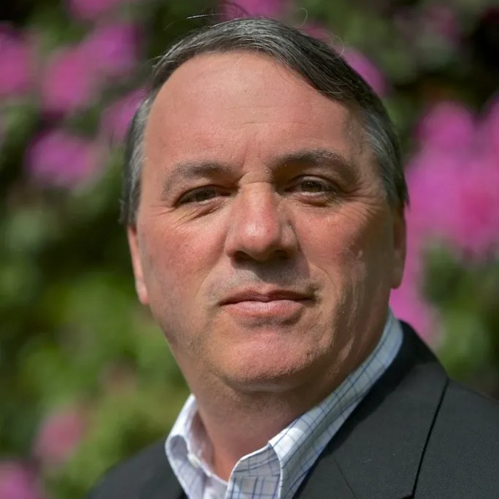 Portrait of a man in a dark blazer and light shirt, standing outdoors with pink flowers blurred behind him