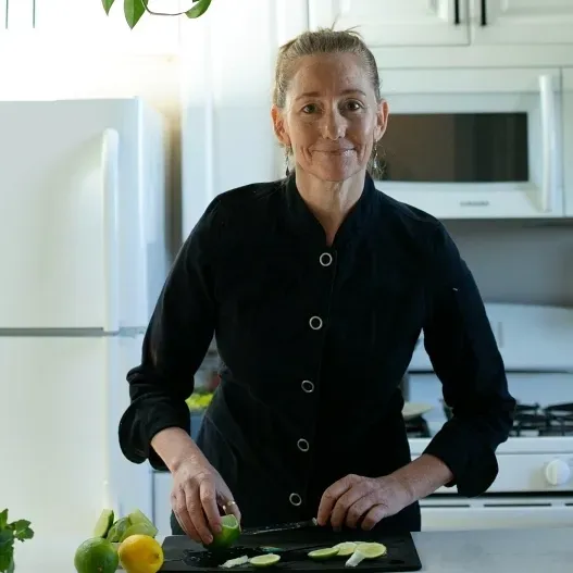 A person in a black chef’s coat slicing limes on a cutting board in a brightly lit kitchen.