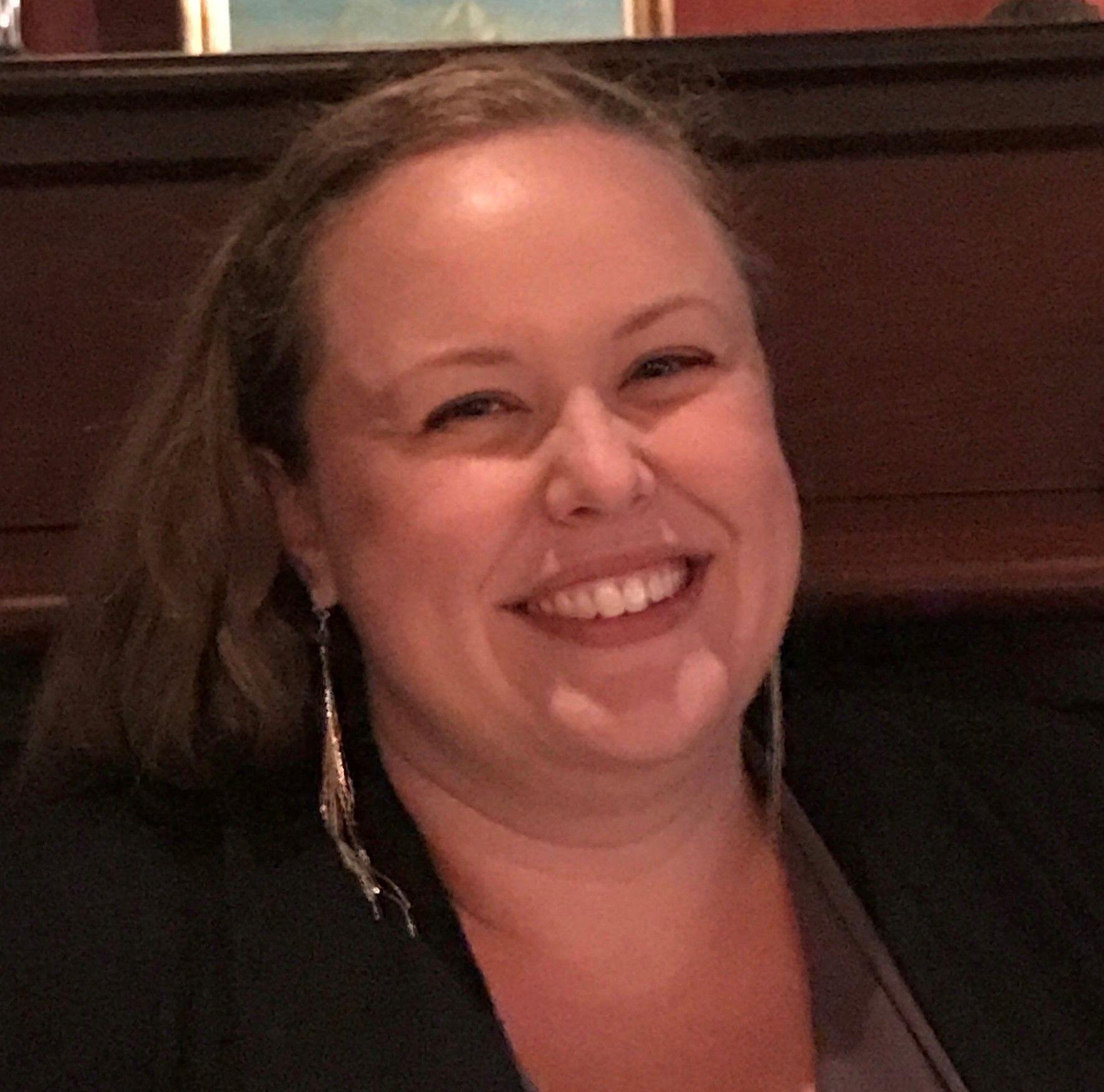A woman wearing a black jacket and earrings smiles for the camera