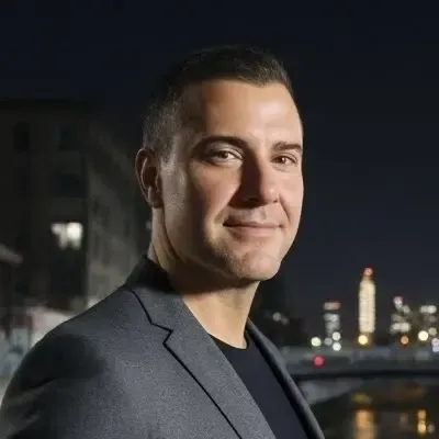 Man with short curly gray hair, wearing earbuds, looking directly at the camera. He wears a dark blue jacket.