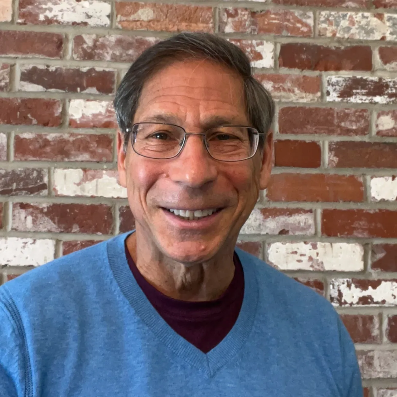 Smiling man in glasses and a blue sweater standing against a brick wall