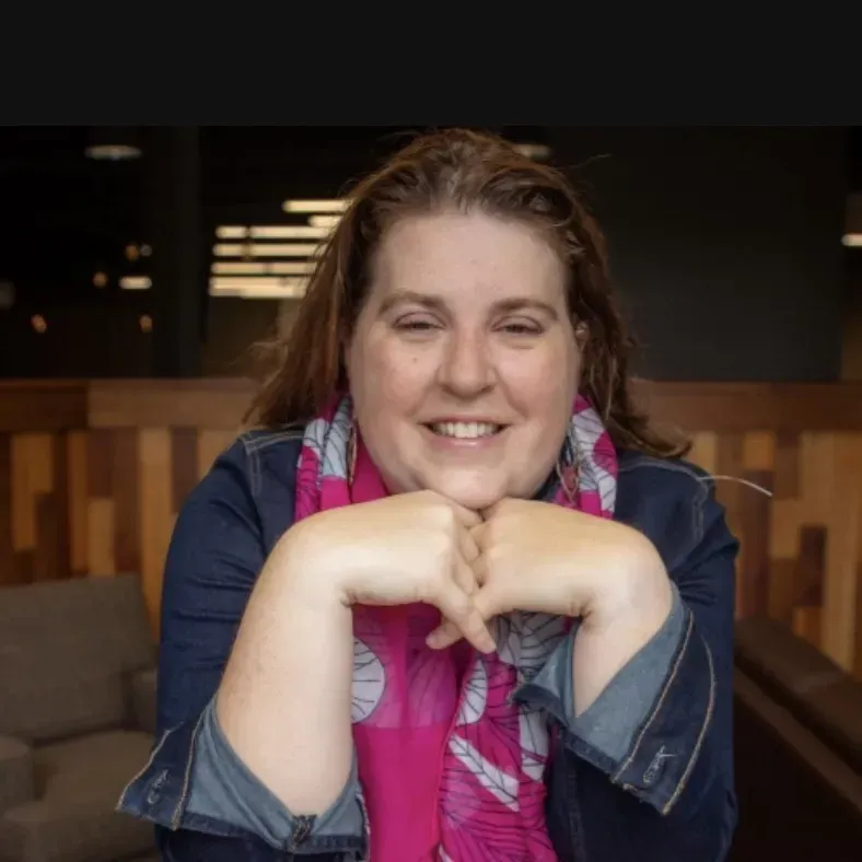 A woman wearing a pink scarf and a denim jacket smiles for the camera