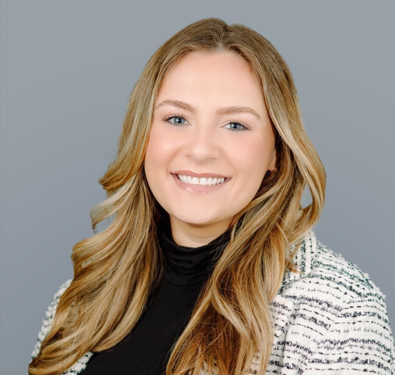A woman with long blonde hair is smiling and wearing a black turtleneck and a white jacket.