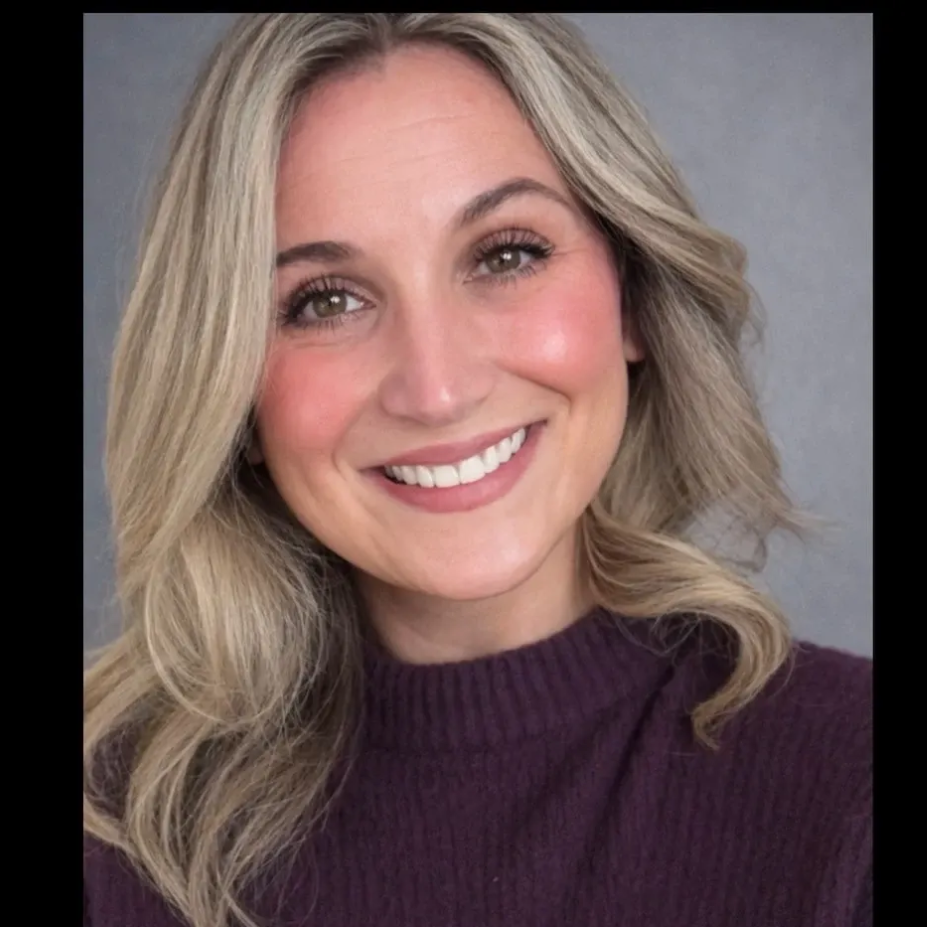 Woman with blonde hair and glasses smiles at the camera, wearing a purple sweater.