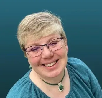 Smiling person with short blond hair and glasses, wearing a teal top and necklace against a blue background