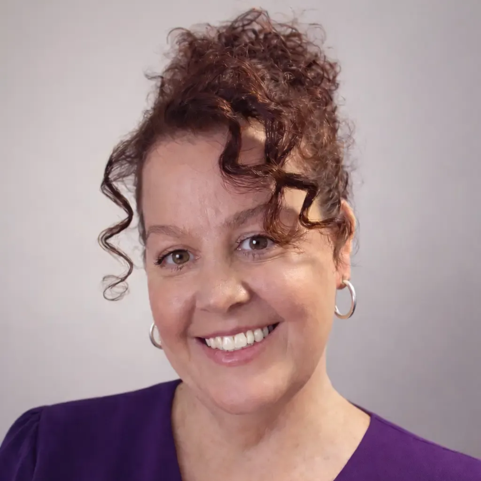 Smiling person with curly hair and hoop earrings wearing a purple top against a light gray background