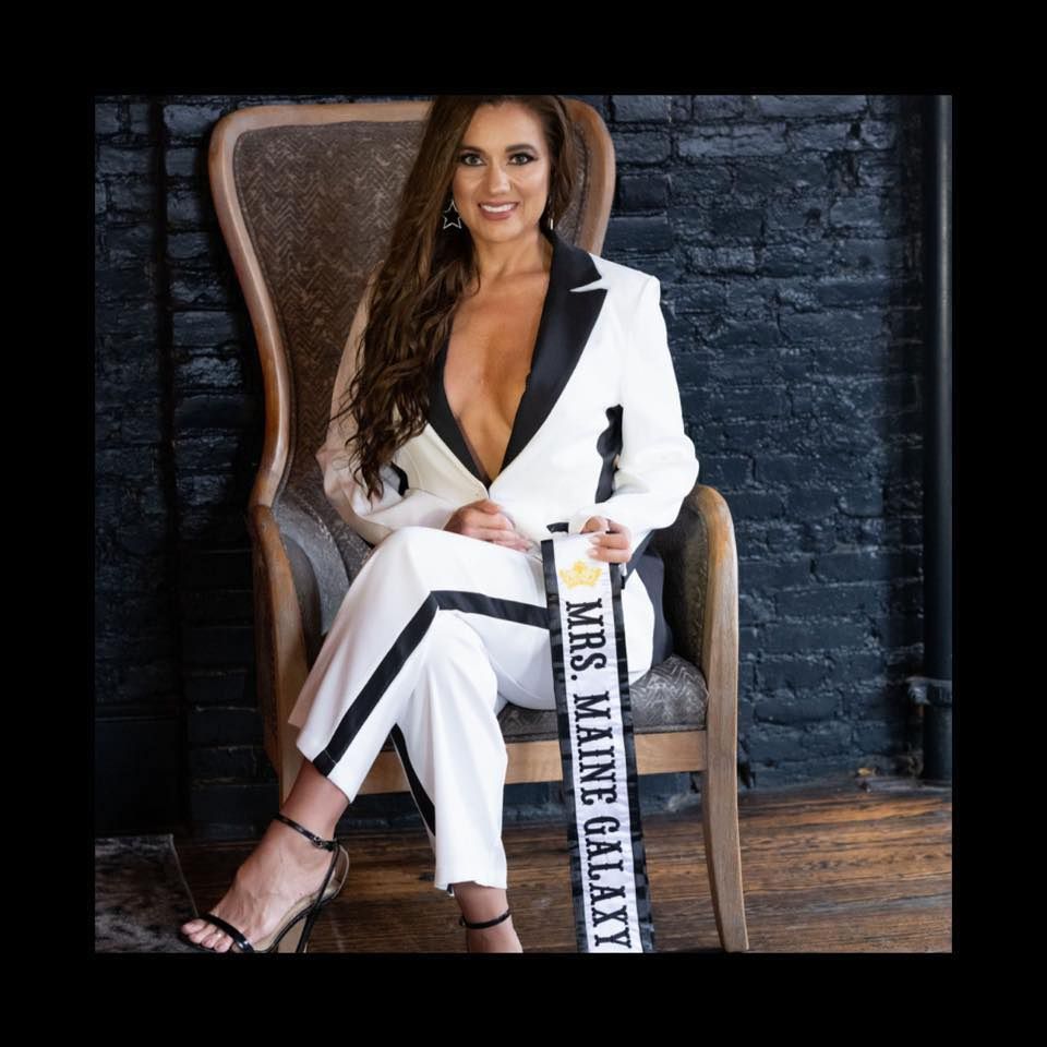 A woman in a suit is sitting in a chair holding a sash that says mrs maine galaxy.