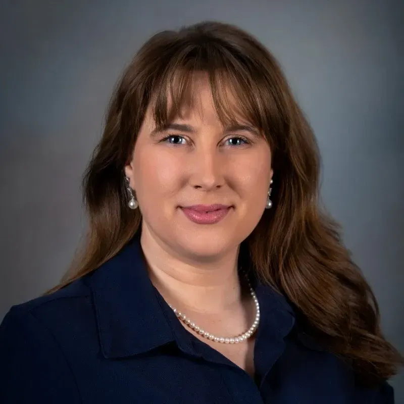 Studio headshot of a woman in a navy blouse, wearing pearl necklace and earrings, against a gray background