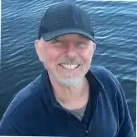 Smiling man in a black cap and navy jacket on a boat, with water behind him