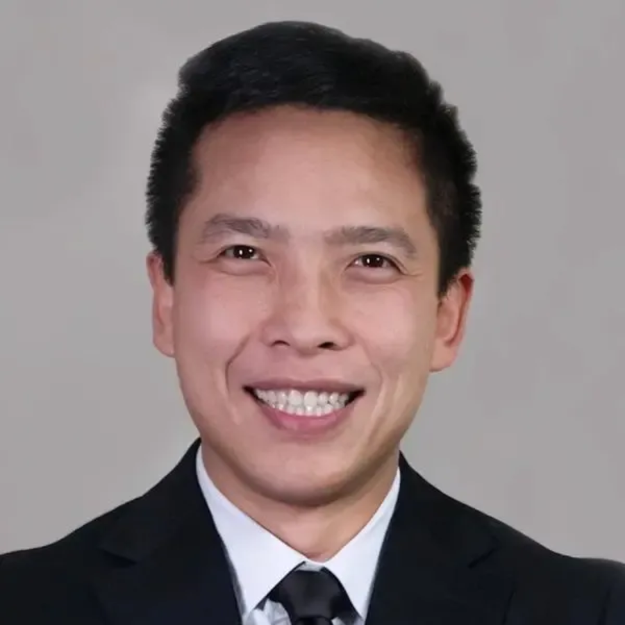 Studio portrait of a smiling person in a black suit, white shirt, and black tie against a gray background