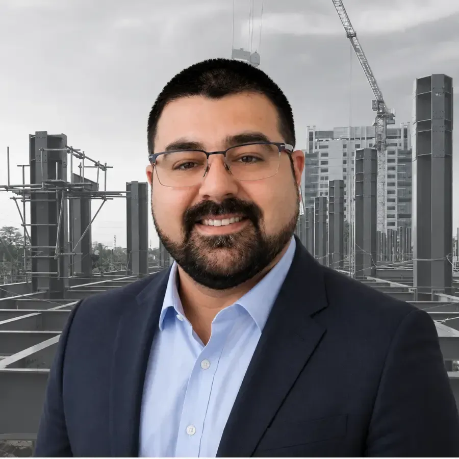 Man in glasses and navy suit smiling at a construction site with cranes and unfinished high-rises behind him