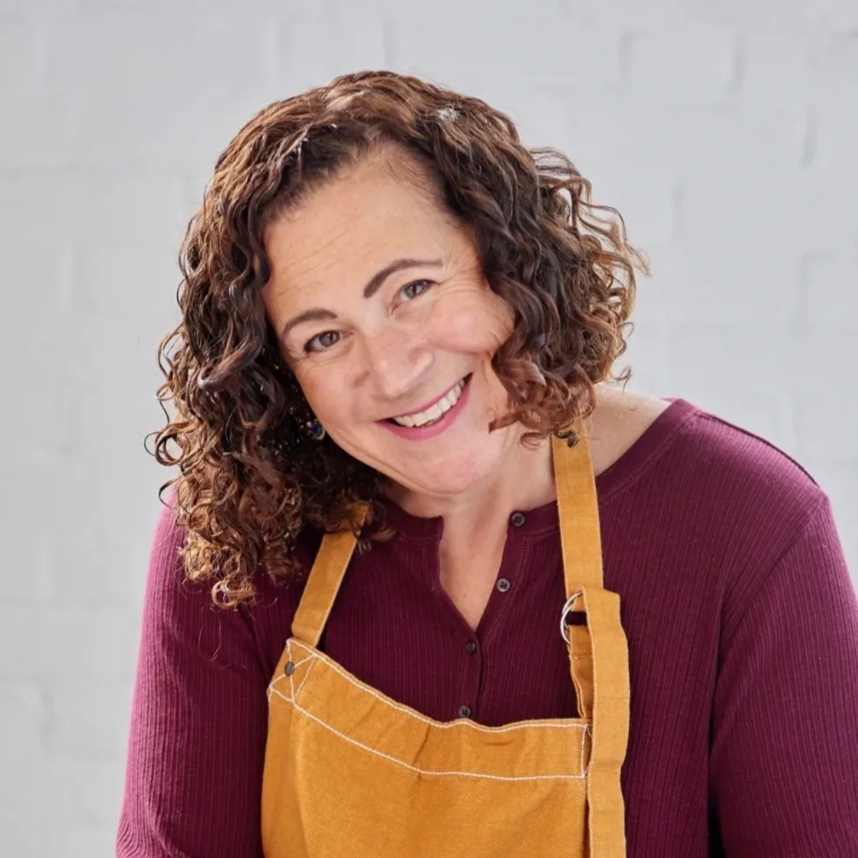 Smiling person with curly hair in a mustard apron and burgundy top against a light gray wall