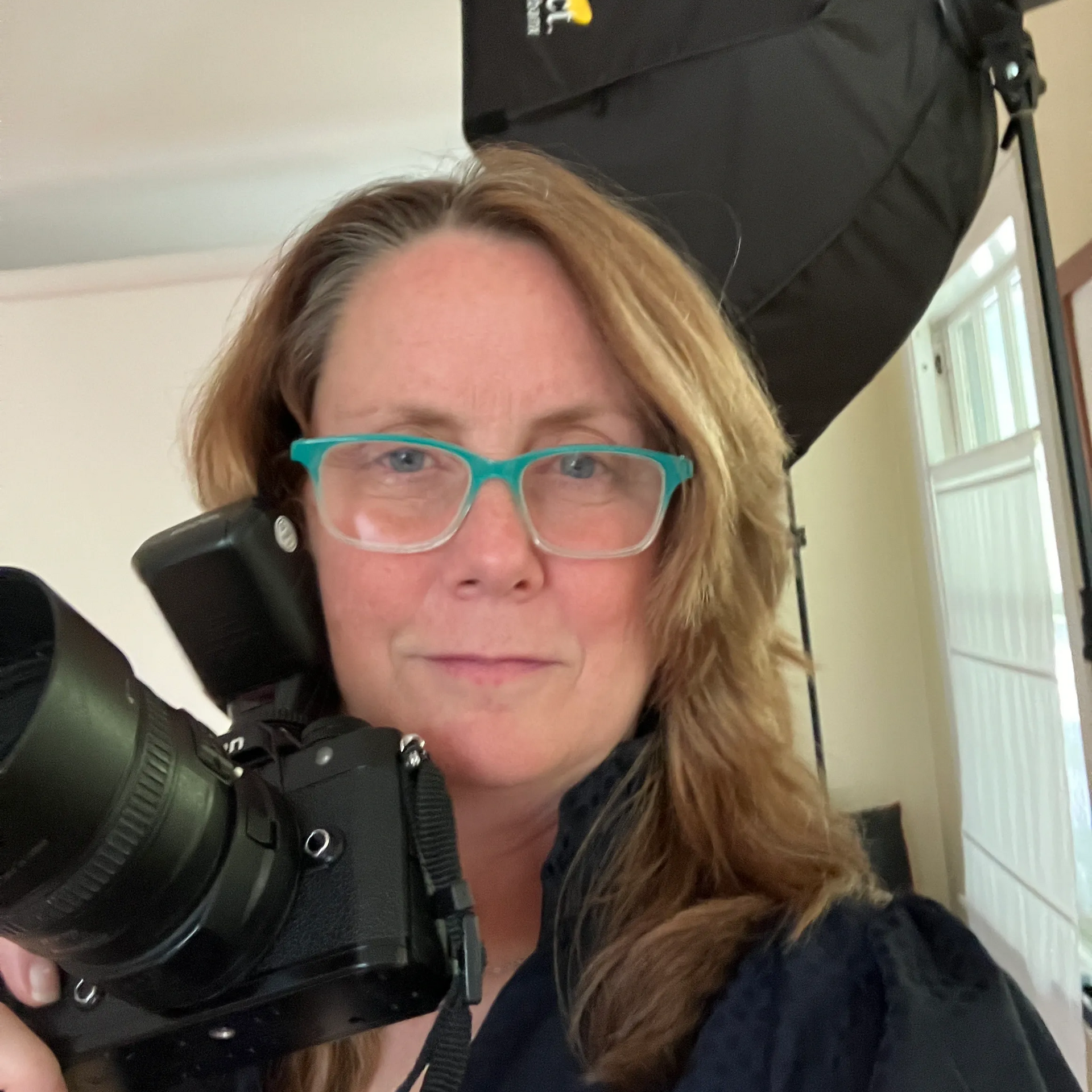 Woman with teal glasses holding a large camera in a studio with a black softbox light behind her