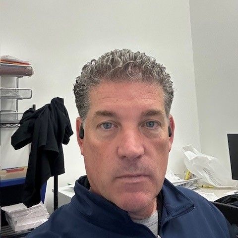 Man with curly gray hair, wearing a dark blue jacket, looking at the camera indoors.