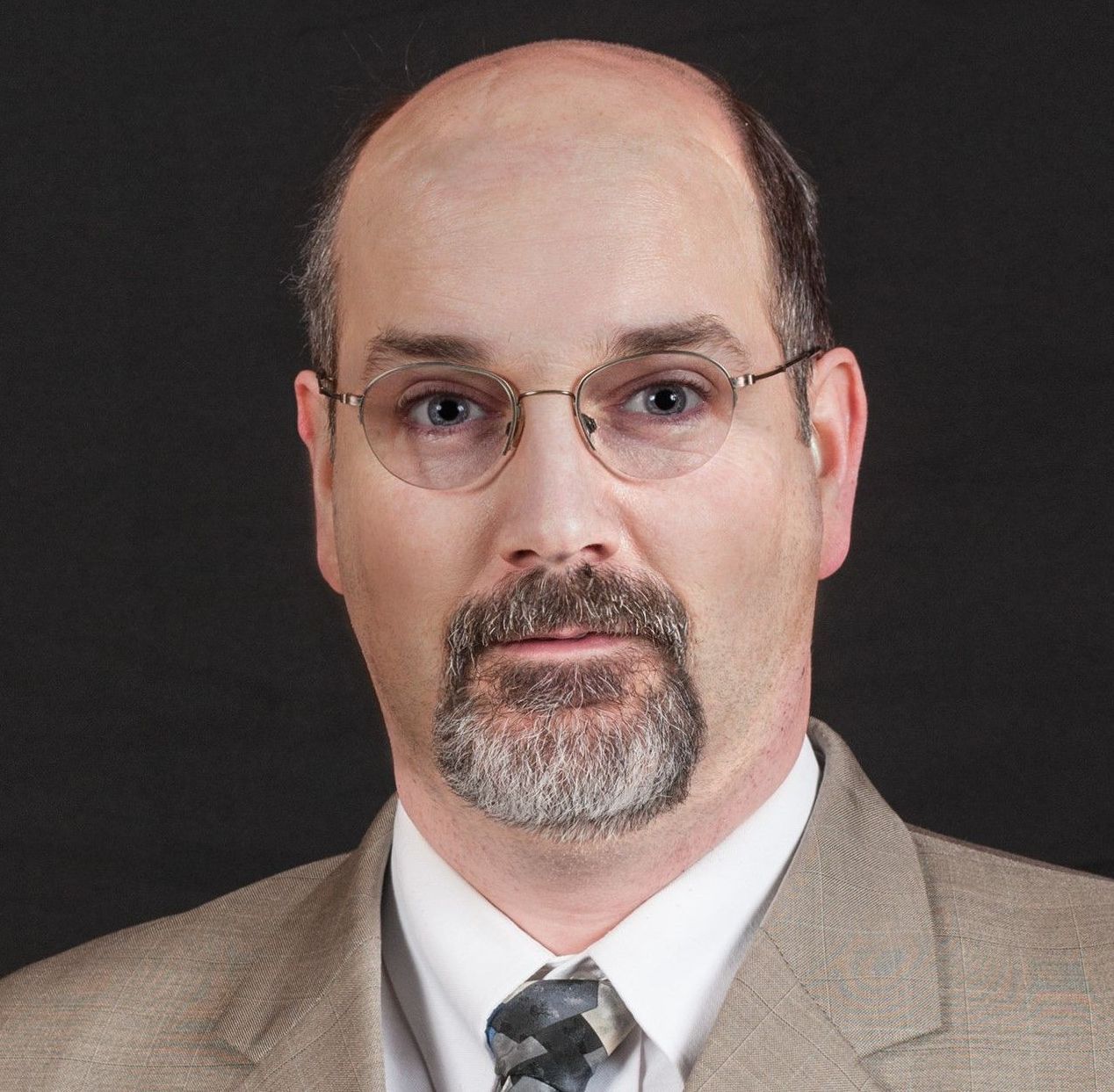 A bald man with glasses and a beard is wearing a suit and tie