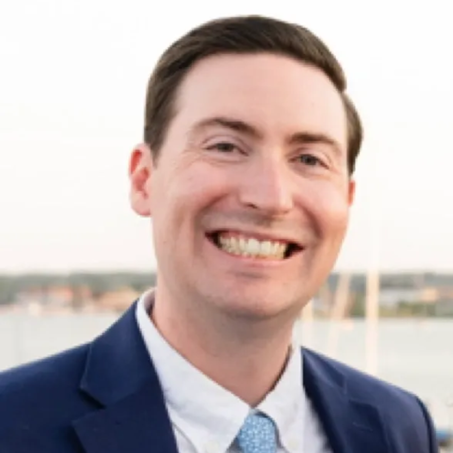 Smiling man in a blue suit outdoors with a blurred waterfront background