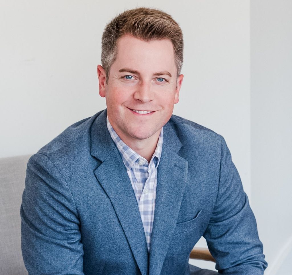 A man in a suit and plaid shirt is sitting in a chair and smiling.