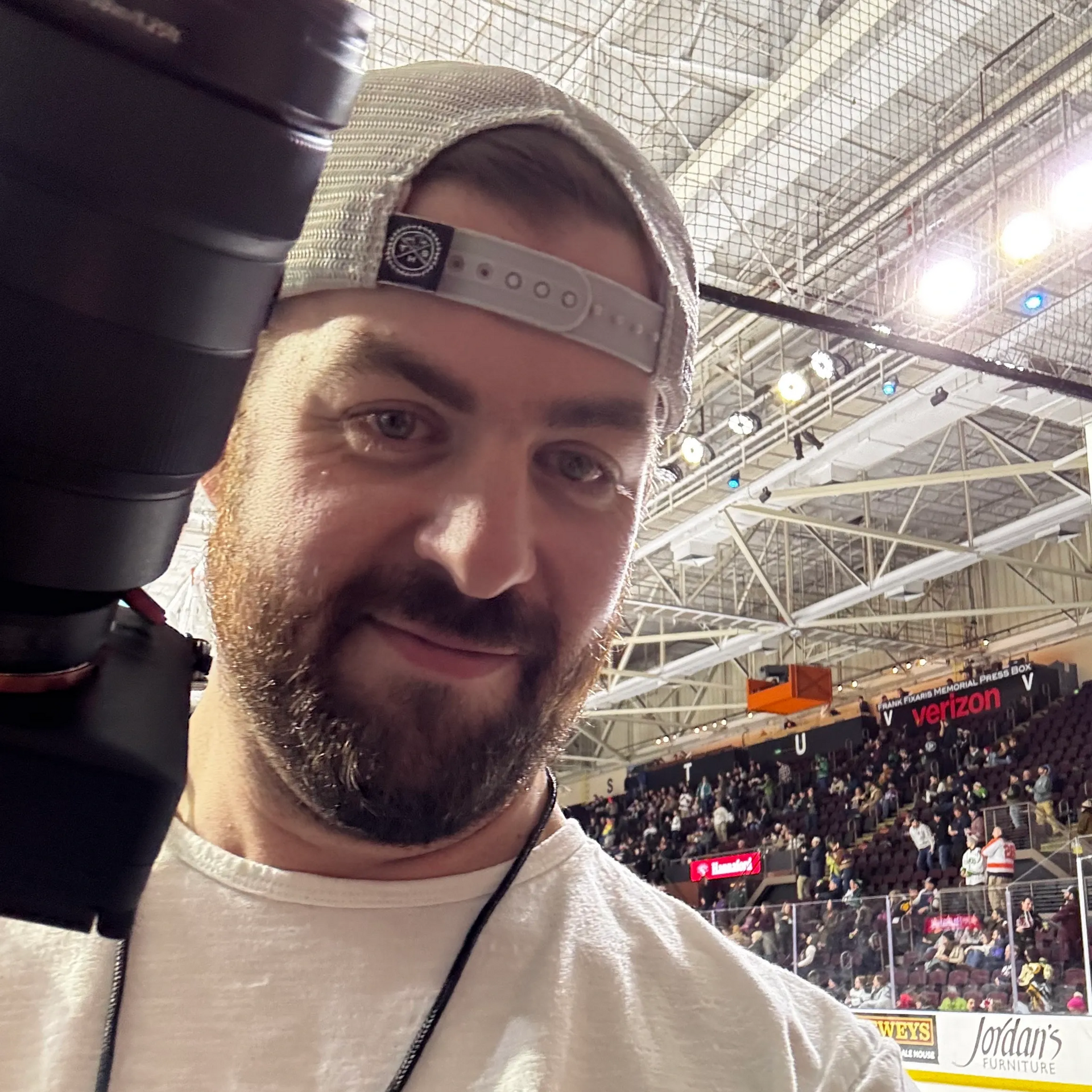 Man smiling in a backward cap at a packed indoor arena, with stadium lights and a hockey rink behind him