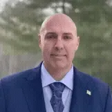Portrait of a bald man in a blue suit and patterned tie, standing outdoors with a blurred green background.