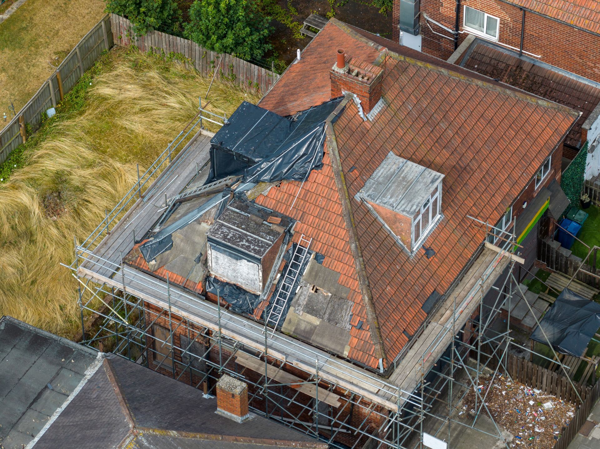 Aerial view of a damaged house roof with tarps and patches, surrounded by fencing and adjacent buildings