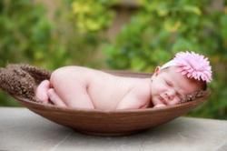 Baby wearing a flower headband, asleep in a brown bowl with a green background.