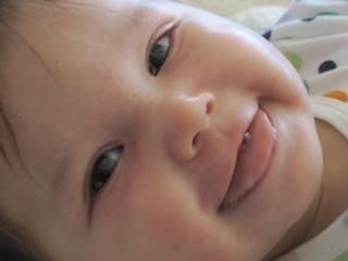 Smiling baby, close-up. Light skin, dark eyes, small teeth showing. Wearing a white shirt.