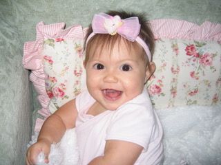 Smiling baby wearing a pink bow headband, sitting in a floral-patterned crib.