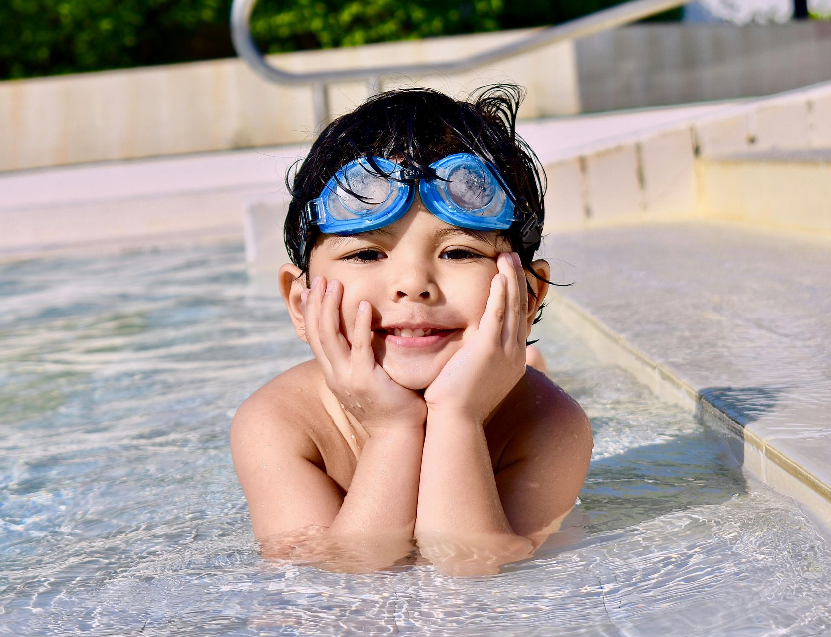 Young boy laying in a shallow pool with goggles on with his hands under his chin.