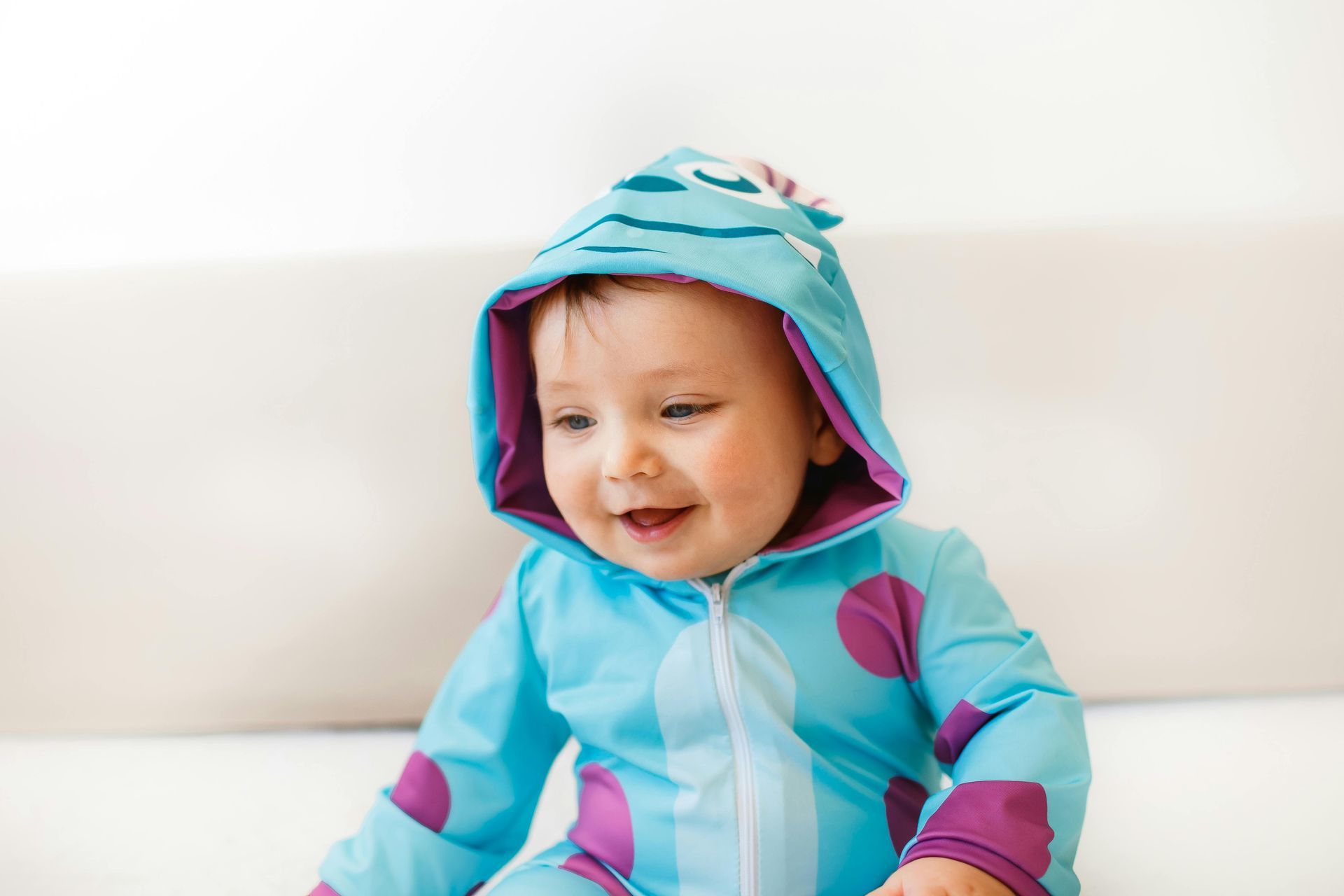 A baby wearing a colorful hoodie is sitting on a white rug and smiling.