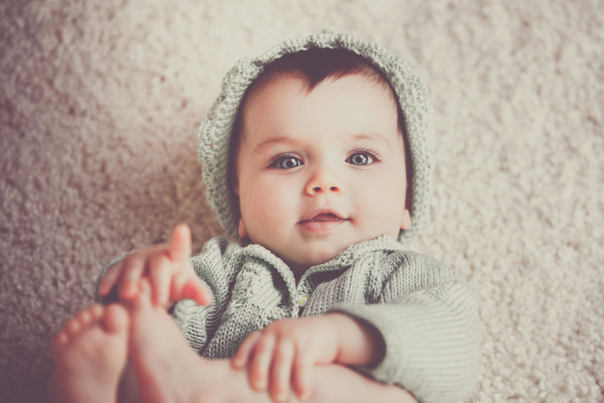 Baby wearing a knitted green hat and sweater, smiling while holding feet, lying on a light-colored rug.