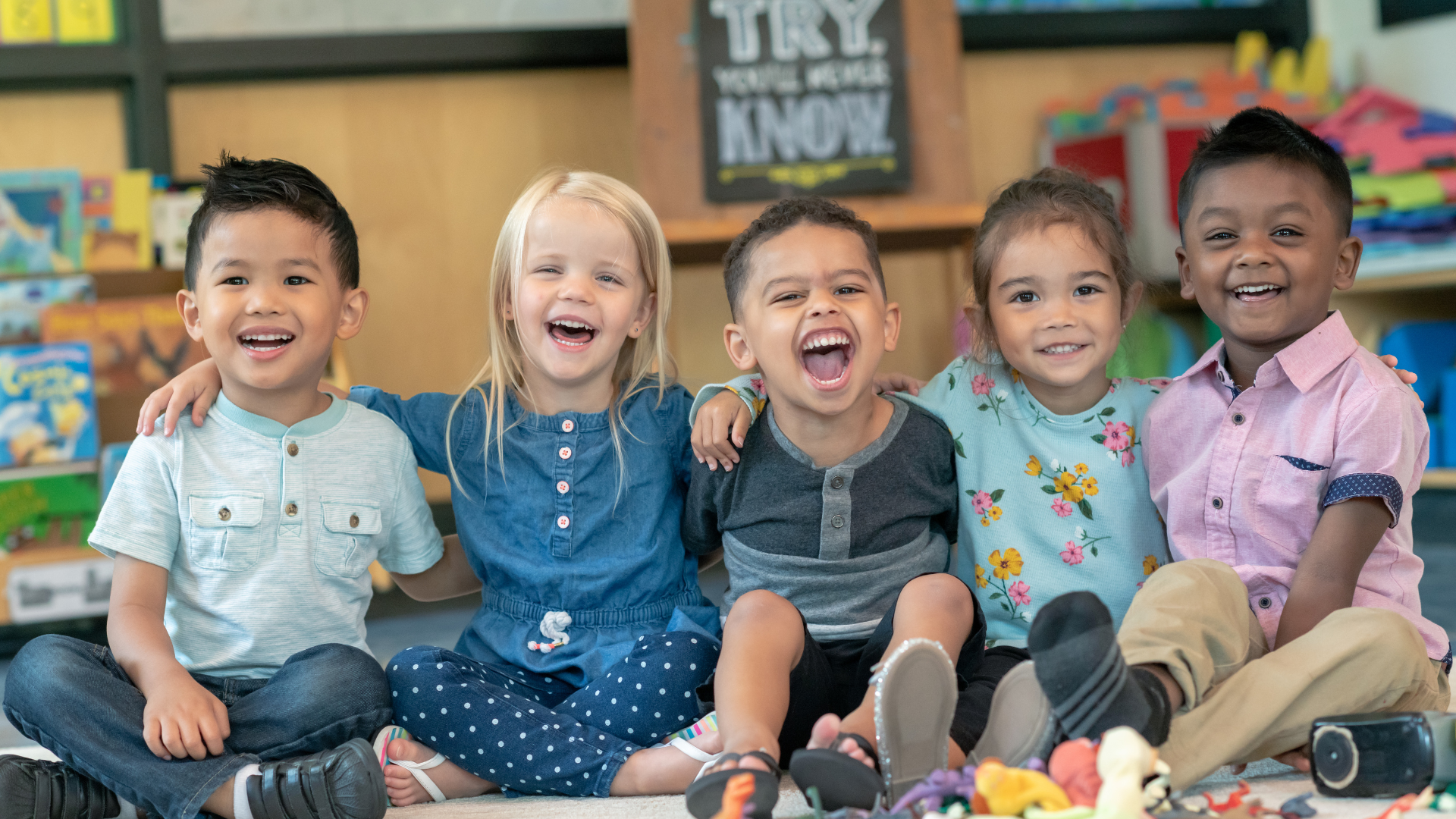 Five diverse children laughing together, sitting on the floor in a classroom.