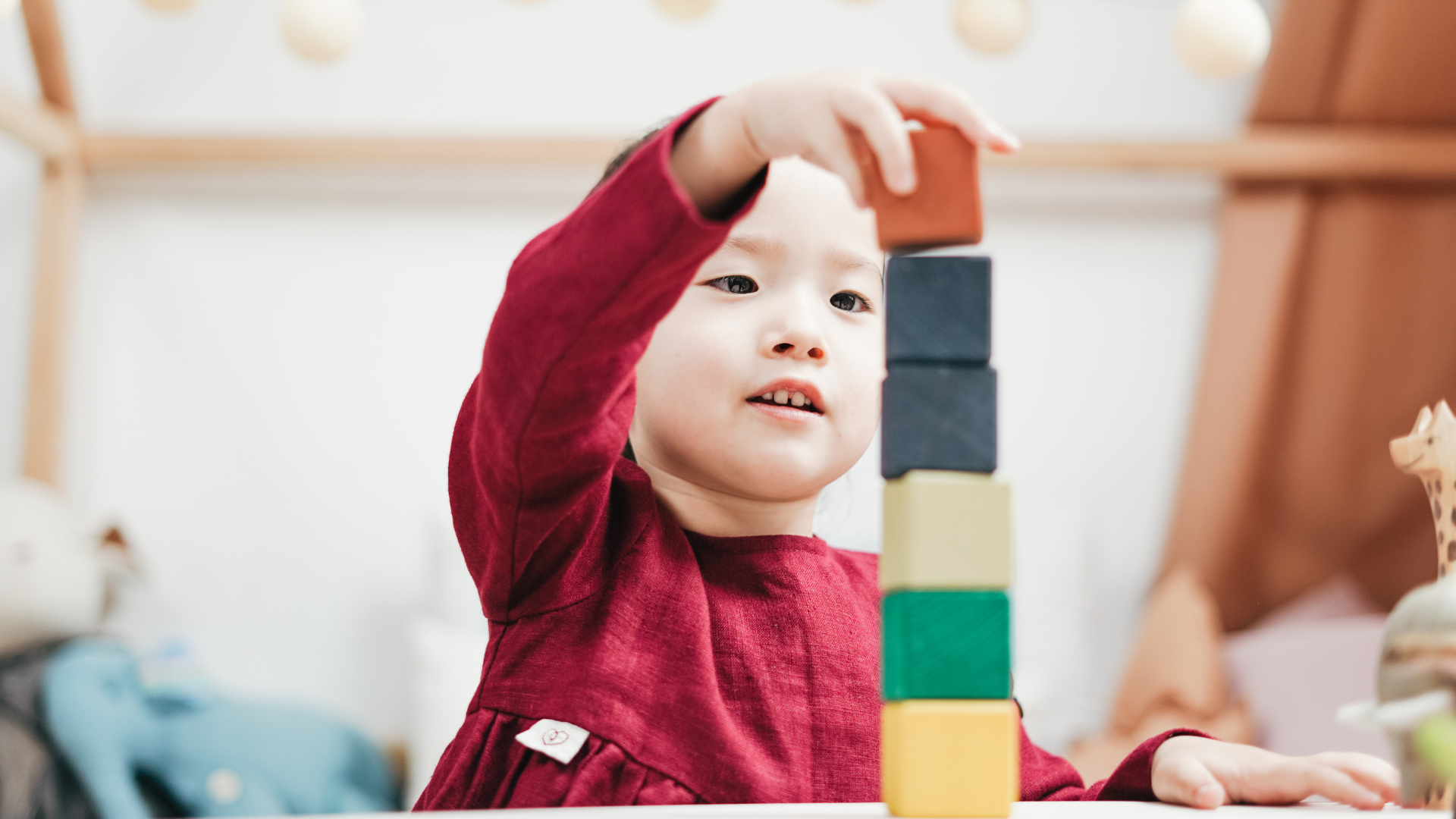 Young child in red dress building a tower of colorful blocks.
