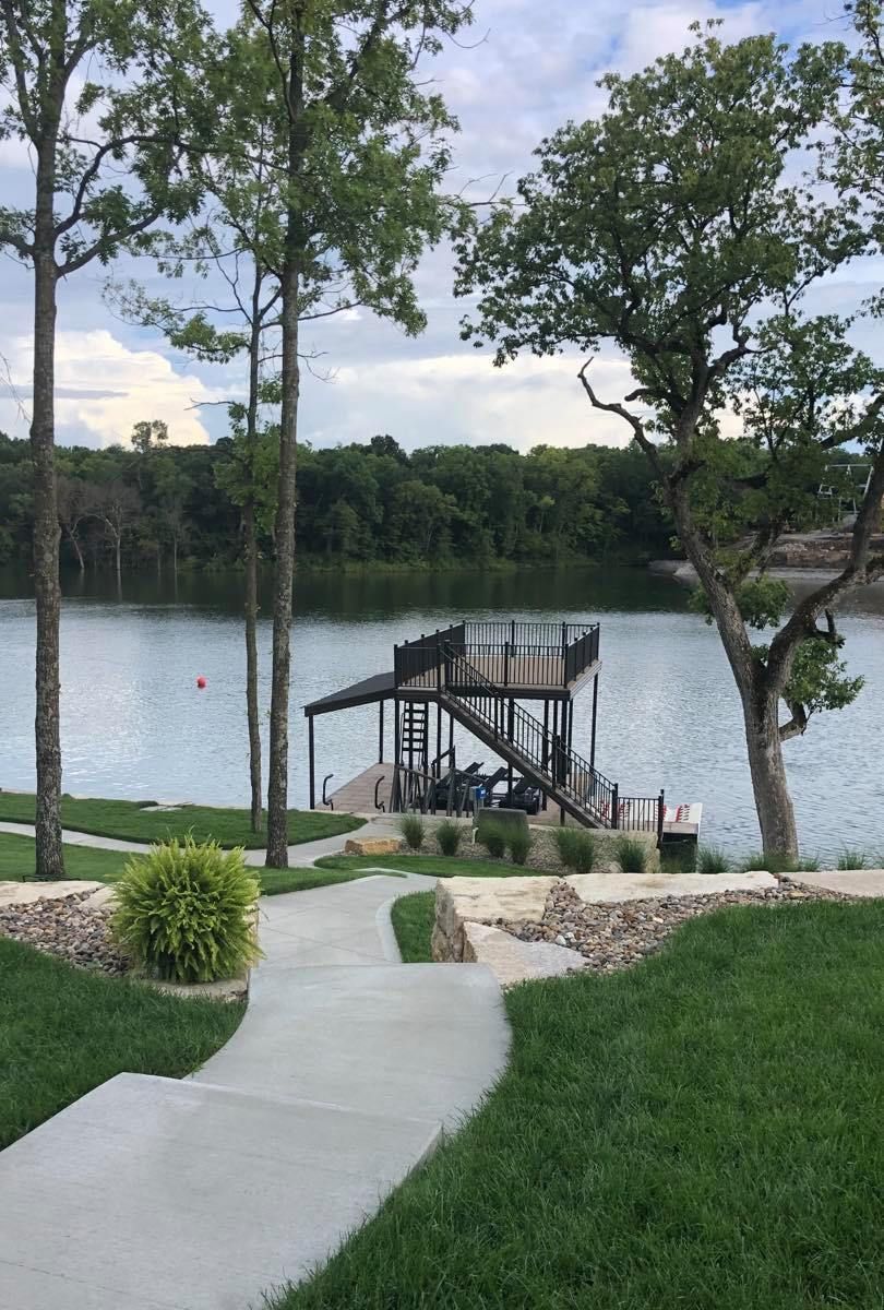 Stone path to a lakeside dock with stairs, surrounded by green lawn and trees under a cloudy sky