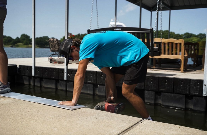 Worker in turquoise shirt crouches on a dock beside a lake, fastening black floating panels.