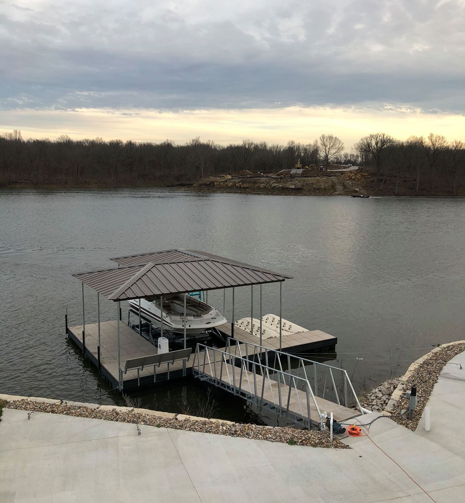 Covered dock on a calm lake beside a concrete shoreline under a cloudy sky.