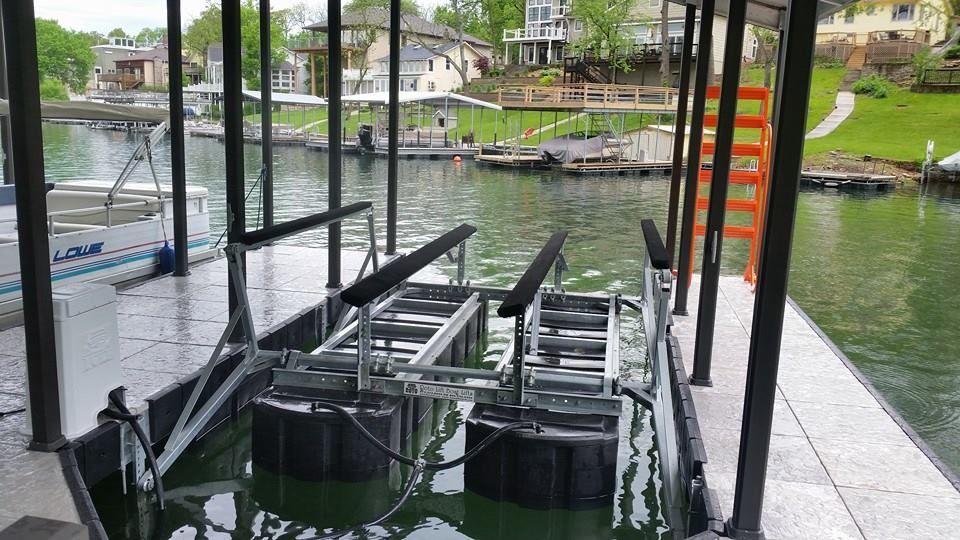 Covered dock with a boat lift beside a calm canal, with houses and a grassy bank in the background