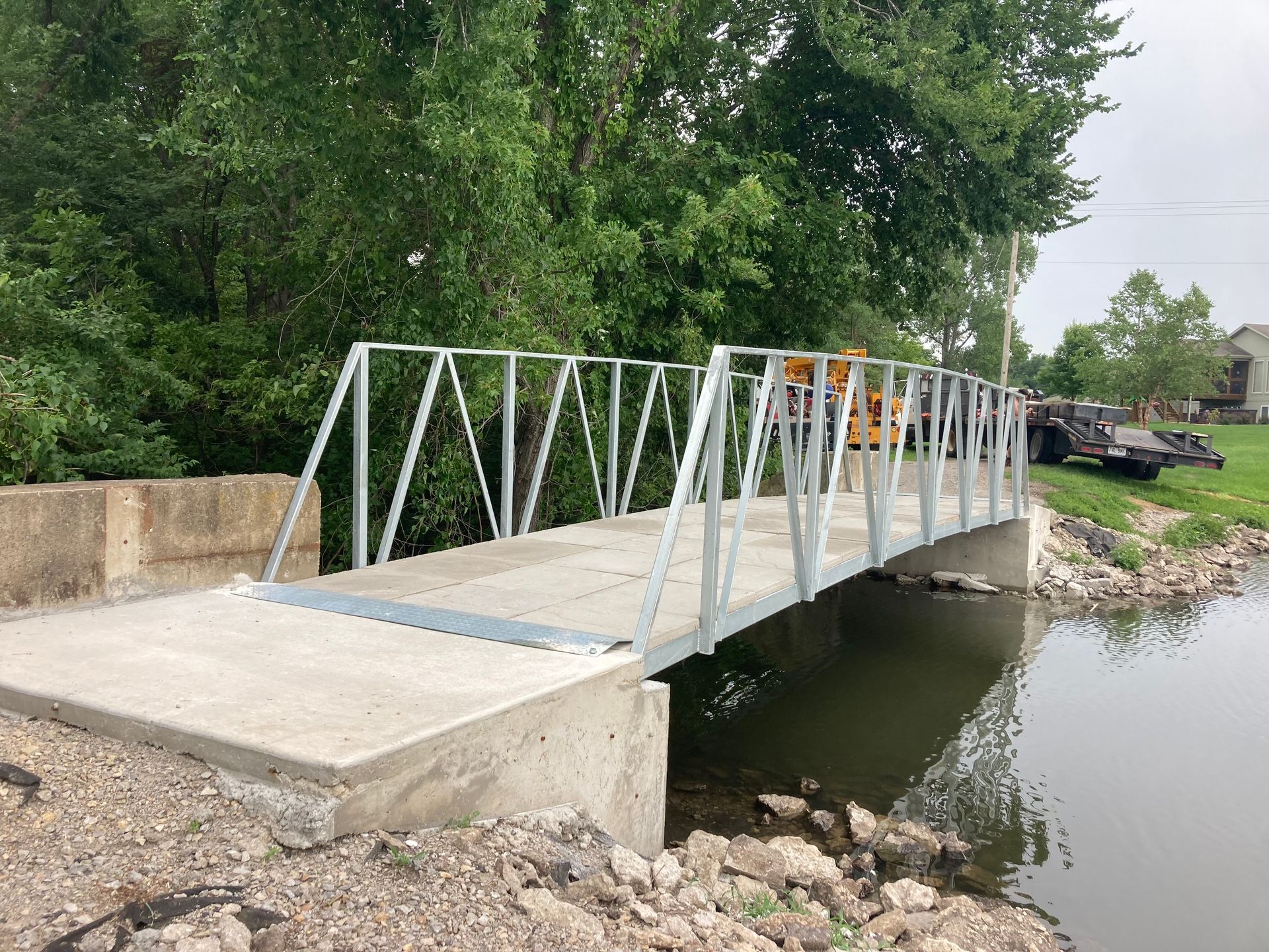 Temporary metal footbridge over a small stream beside a concrete abutment and green trees