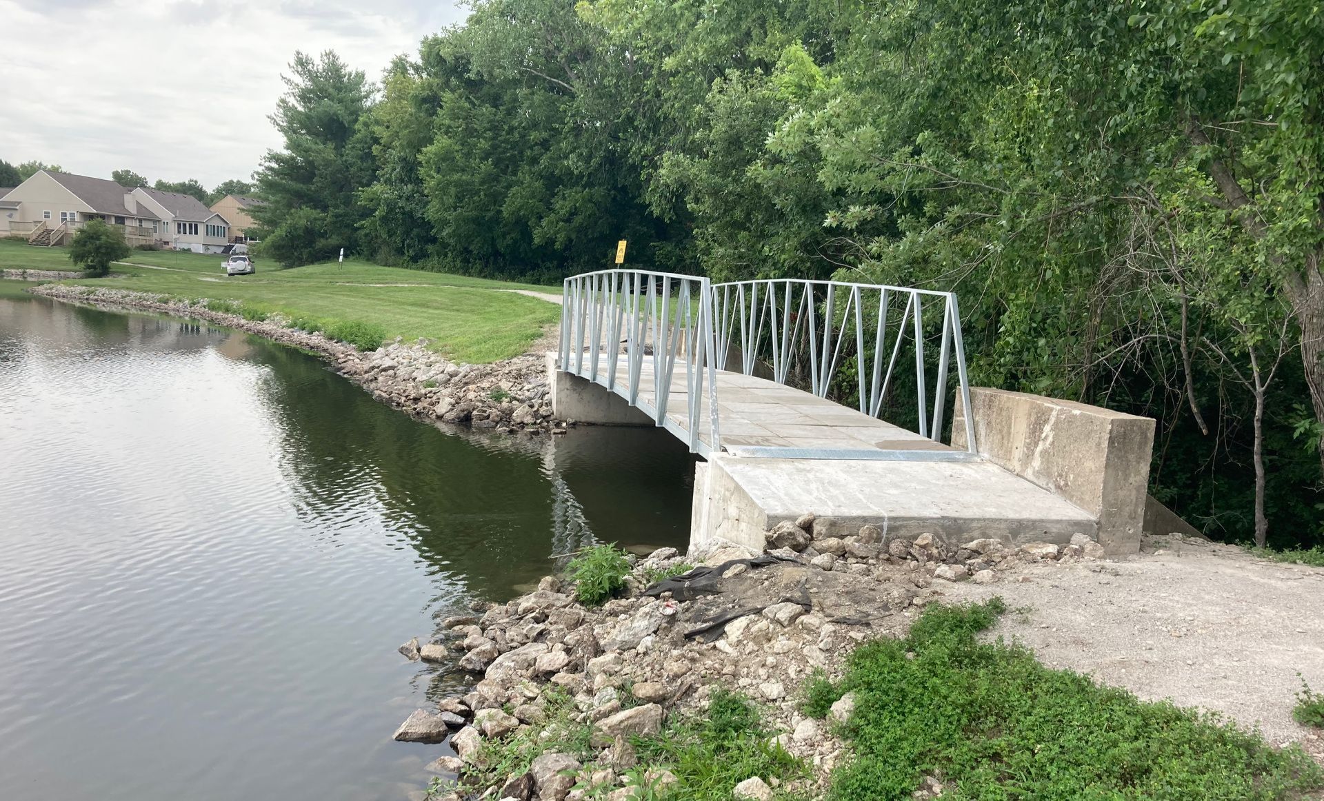 Small metal footbridge over a calm pond, bordered by rocks and trees in a park.