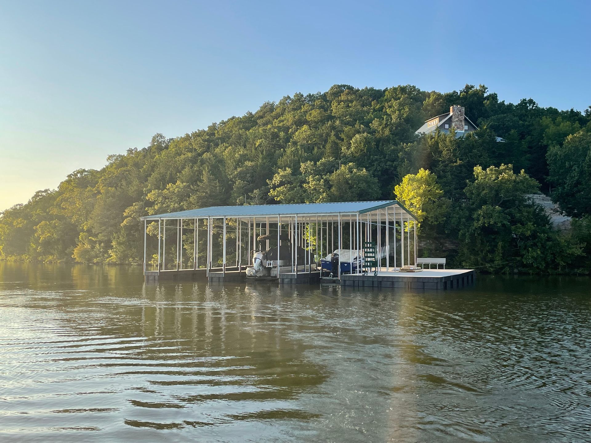 Lakefront dock with a covered seating area, backed by a wooded hillside at sunset